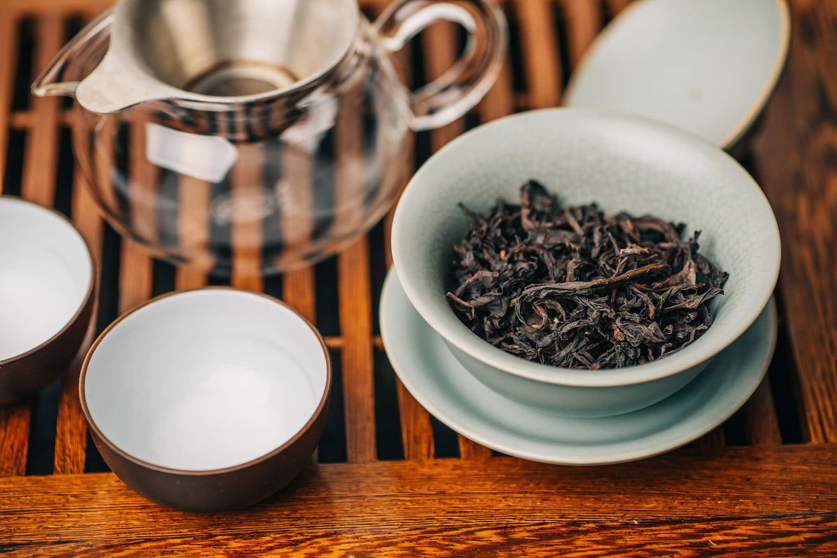 A traditional tea setup featuring a bowl of loose-leaf Da Hong Pao oolong tea, a glass teapot with a metal strainer, and two small teacups on a wooden tea tray, capturing the essence of Chinese tea culture.