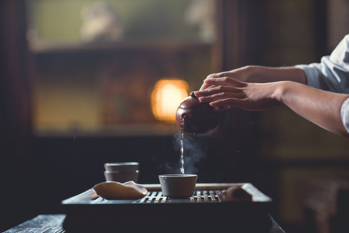 An individual gracefully pours steaming Chinese tea from a traditional teapot into a teacup, set against a warm, ambient background. The scene captures a moment of tranquility and elegance in a Chinese tea ceremony.