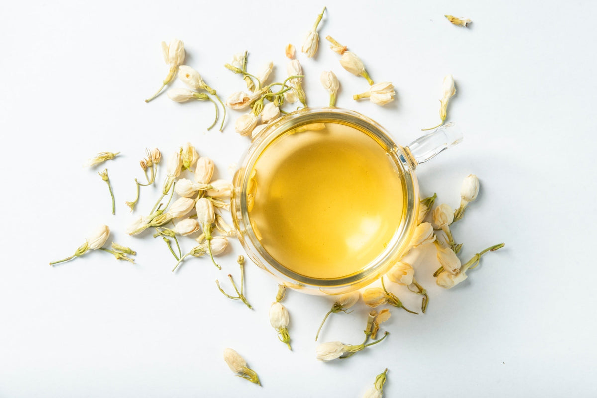 A glass cup of freshly brewed jasmine tea with a golden hue, surrounded by delicate dried jasmine flowers on a white background.