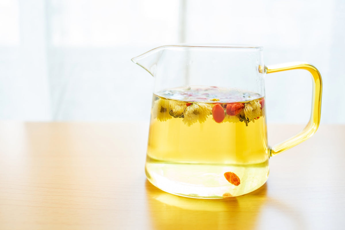 A clear glass teapot filled with golden chrysanthemum tea, with dried chrysanthemum flowers and goji berries floating on the surface. The teapot sits on a wooden table with a bright, airy background, creating a refreshing and calming tea-drinking ambianc