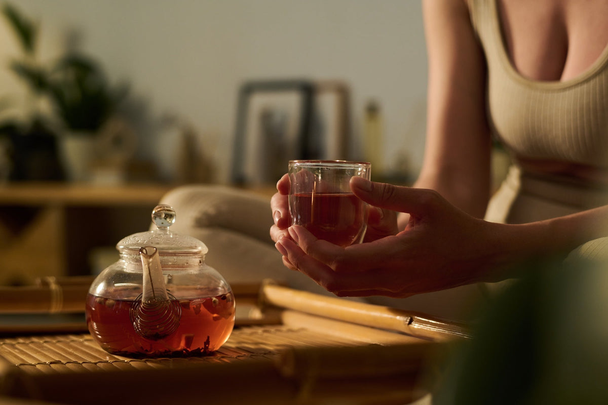 A woman enjoying a warm cup of freshly brewed tea in a cozy, relaxed setting, with a glass teapot steeping loose leaf tea on a bamboo tray.