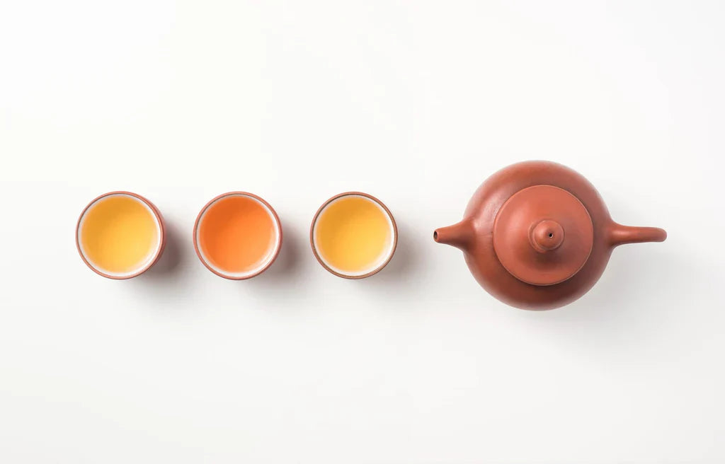This image features a minimalist arrangement of a traditional Chinese teapot and three teacups displaying a gradient of tea colors, from light yellow to a deep amber, symbolizing different tea varieties or steeping levels. The setup showcases the elegance