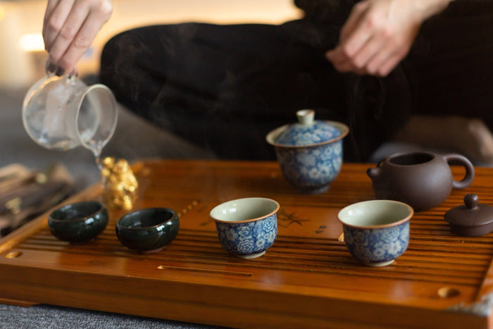 A traditional Chinese tea ceremony setup with a wooden tea tray, porcelain teacups, and a Yixing clay teapot. Hot water is being poured into a cup, showcasing the authentic way to properly brew tea.