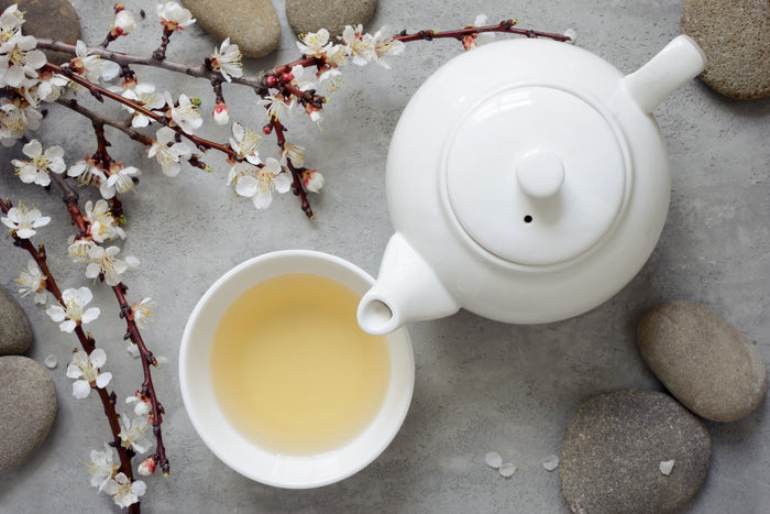 A white porcelain teapot and teacup filled with delicate White Peony tea, accompanied by blooming white flowers and smooth stones on a textured gray surface, evoking a sense of tranquility and elegance.