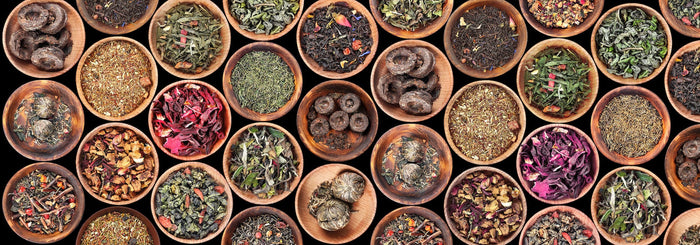 A diverse selection of loose-leaf teas displayed in wooden bowls, showcasing various tea types such as green tea, black tea, oolong tea, white tea, herbal blends, and compressed Pu-erh tea.