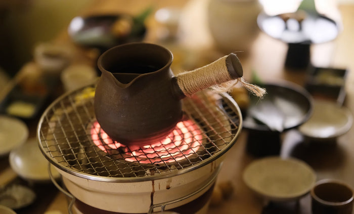 A traditional ceramic teapot heating over an open flame on a stove with a wire mesh. The rustic tea-making setup is surrounded by teaware and utensils, creating a warm and authentic tea-brewing atmosphere.