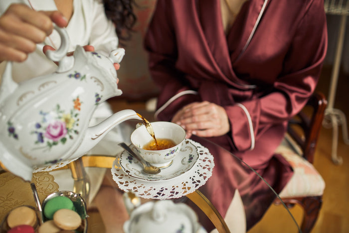 A luxurious afternoon tea setting with two elegantly dressed women enjoying tea served from a floral porcelain teapot, accompanied by delicate macarons on a glass table.