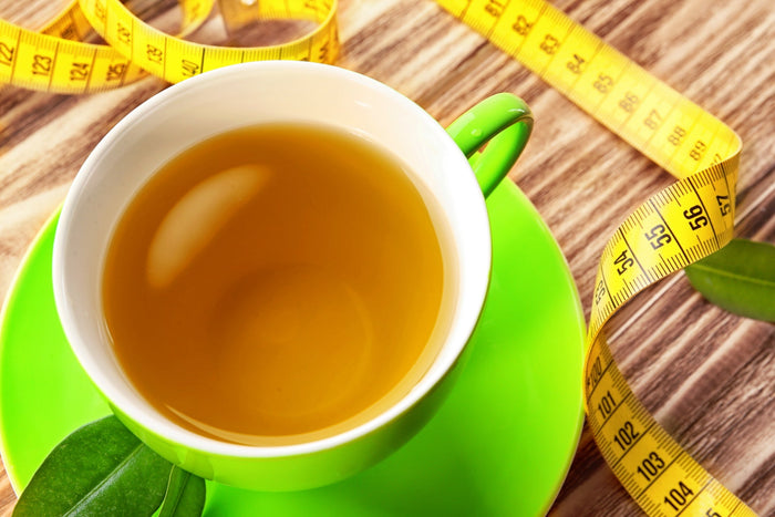 A cup of green tea in a vibrant green cup and saucer, placed on a wooden surface with fresh tea leaves and a yellow measuring tape, symbolizing weight loss and metabolism-boosting benefits of tea.
