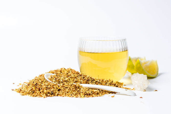 An elegant display of Osmanthus tea featuring a pile of dried osmanthus flowers beside a glass of freshly brewed tea, showcasing its light golden hue. In the background, lime wedges and a crystal add a refreshing touch.