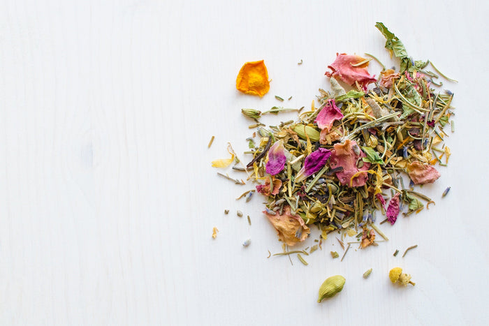 A colorful blend of dried herbs, rose petals, chamomile flowers, and other botanicals on a white wooden surface, representing a floral herbal tea mix.