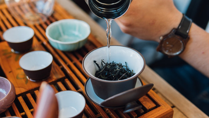 A close-up of traditional Chinese oolong tea brewing using a gaiwan. Hot water is being poured over loose-leaf oolong tea, with small teacups and a wooden tea tray in the background.