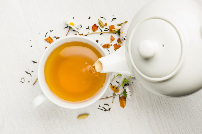 A white ceramic teapot pouring golden herbal tea into a matching cup, surrounded by loose tea leaves, dried flowers, and herbs on a white wooden surface. The scene highlights the freshness and natural ingredients of organic loose leaf tea.