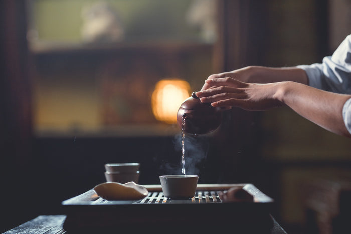 An individual gracefully pours steaming Chinese tea from a traditional teapot into a teacup, set against a warm, ambient background. The scene captures a moment of tranquility and elegance in a Chinese tea ceremony.