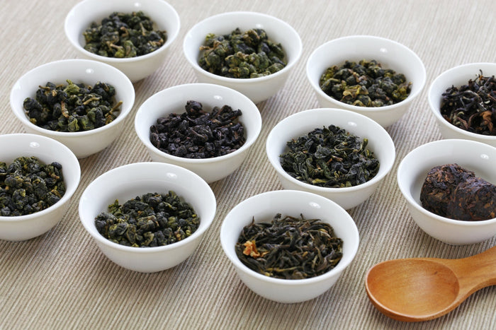 An assortment of Chinese oolong tea varieties displayed in small white bowls, showcasing different leaf shapes, oxidation levels, and processing styles on a textured fabric surface.
