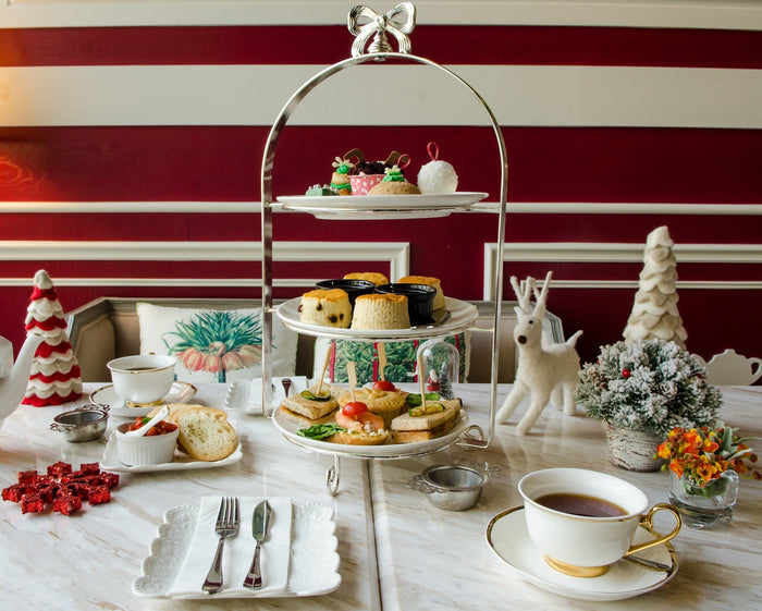 A beautifully arranged afternoon tea setup for Mother's Day, featuring a three-tiered stand with pastries, scones, and sandwiches. The table is elegantly decorated with teacups, a teapot, festive holiday ornaments, and floral arrangements, creating a cozy
