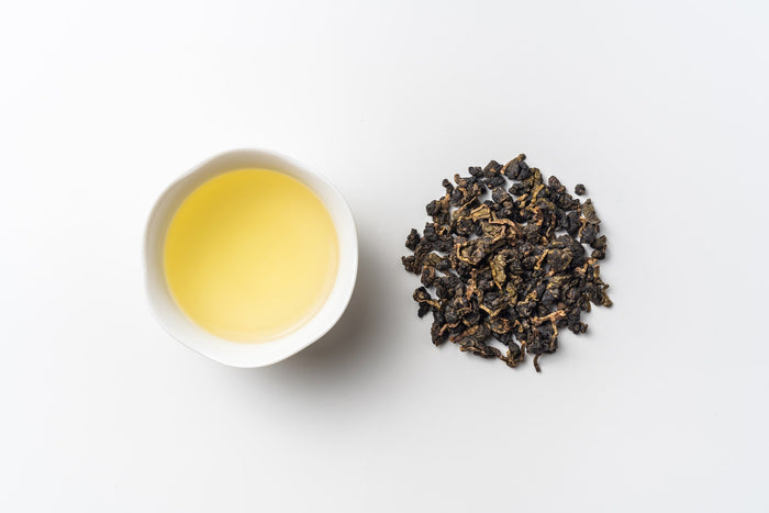 A white ceramic cup filled with freshly brewed roasted oolong tea with a golden hue, placed next to a pile of tightly rolled roasted oolong tea leaves on a clean white background.