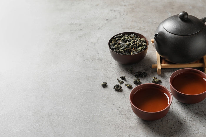 A traditional tea setup featuring a black teapot, a bowl of loose-leaf rolled oolong tea, and two cups of freshly brewed amber-colored oolong tea on a textured gray surface.