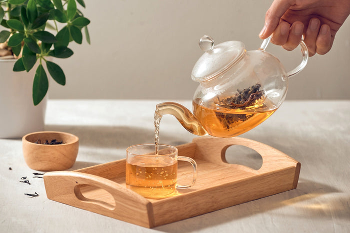 A glass teapot filled with freshly steeped oolong tea being poured into a clear glass cup, placed on a wooden tray. A bowl of loose oolong tea leaves sits nearby, adding to the serene tea experience.