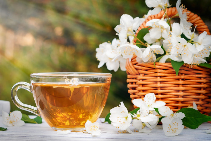 A glass cup of golden jasmine tea on a white wooden surface, accompanied by fresh jasmine flowers and a wicker basket filled with blooming jasmine blossoms, set against a sunlit green garden background.