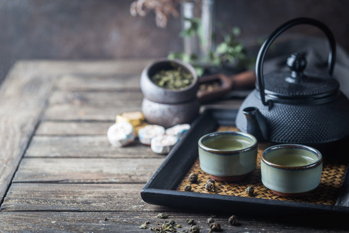 A rustic tea setup featuring a traditional black cast iron teapot, two ceramic cups filled with green tea, and loose tea leaves on a wooden tray. The background includes a stone bowl with dried tea leaves and compressed tea cakes, creating an authentic te