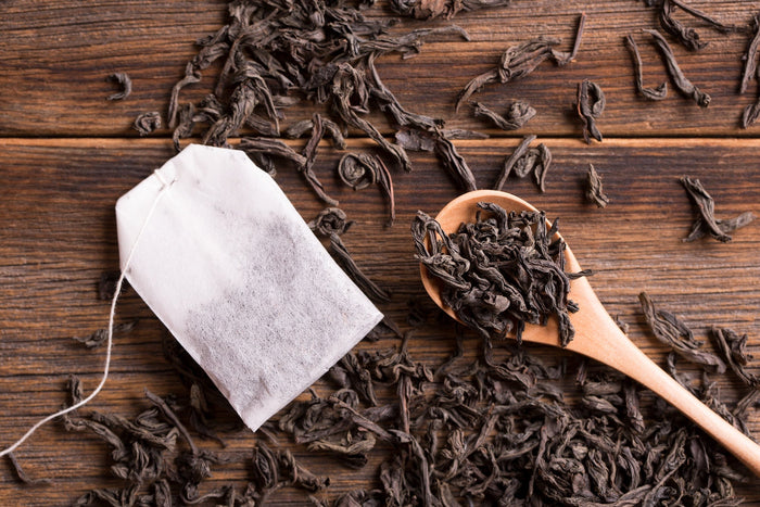 A close-up of an organic black tea bag placed on a rustic wooden surface, accompanied by a wooden spoon filled with loose black tea leaves. The rich, dried tea leaves emphasize the quality and authenticity of premium organic black tea.