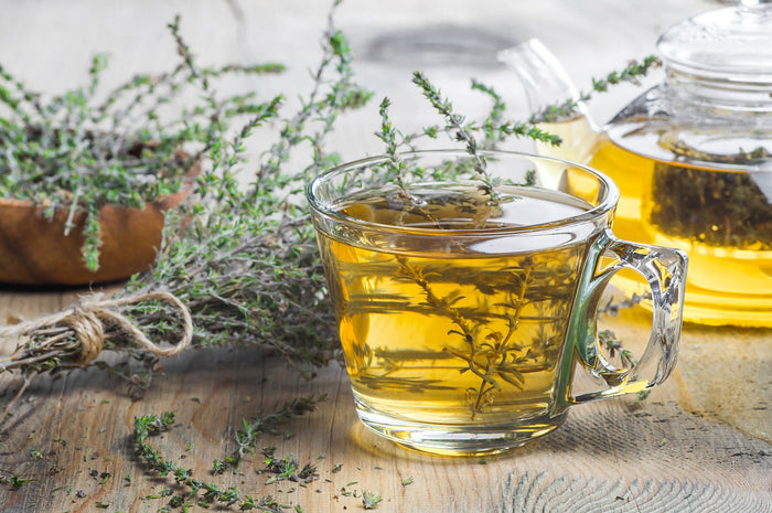 A steaming cup of herbal green tea infused with fresh thyme, placed on a rustic wooden table with a glass teapot and a bundle of thyme in the background.