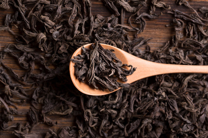 A close-up of loose-leaf black tea on a wooden spoon, surrounded by dried tea leaves on a rustic wooden surface. The dark, twisted tea leaves highlight the rich texture and quality of premium black tea, commonly used in traditional tea brewing.