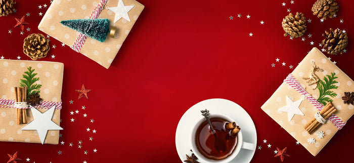 A festive Christmas tea scene with a cup of spiced tea garnished with cinnamon and star anise, surrounded by beautifully wrapped holiday tea gift sets. The red background, pinecones, and sparkling decorations create a warm and cozy holiday atmosphere, per