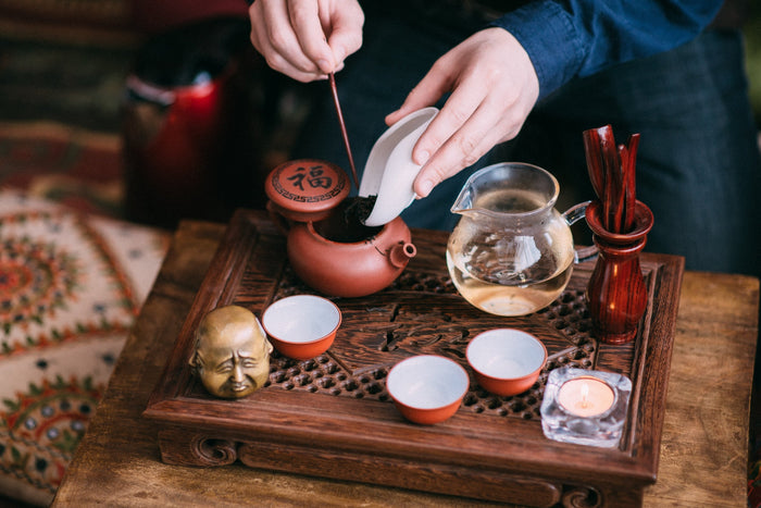 Person preparing tea during a traditional Chinese tea ceremony with a clay teapot, glass pitcher, and small teacups on a carved wooden tray.
