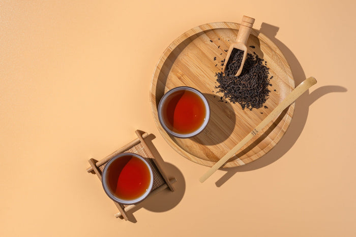 Two cups of brewed black tea placed on a wooden tray with loose tea leaves and a bamboo scoop, set against a warm beige background.