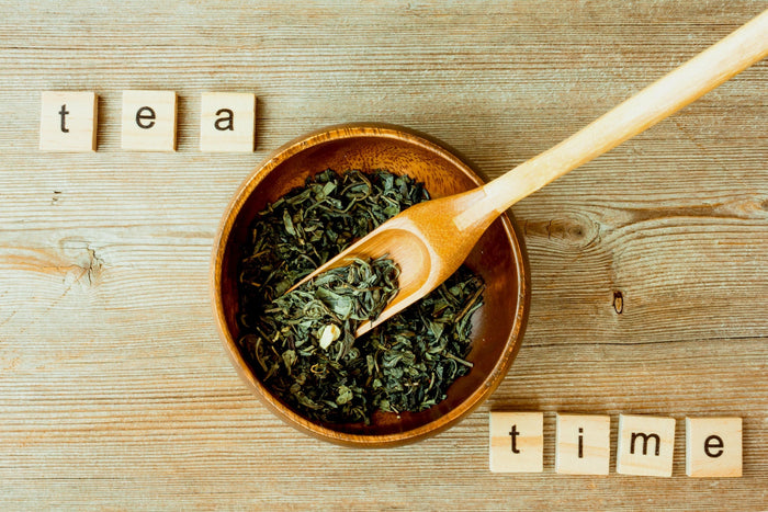 A wooden bowl filled with loose-leaf tea on a rustic wooden table, with a wooden scoop resting inside. Surrounding the bowl, small wooden letter tiles spell out 'tea time' in a cozy, inviting arrangement.