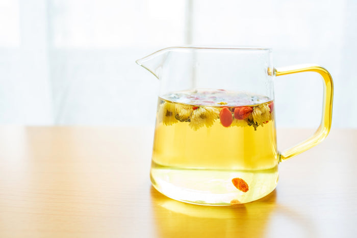  A clear glass teapot filled with golden chrysanthemum tea, with dried chrysanthemum flowers and goji berries floating on the surface. The teapot sits on a wooden table with a bright, airy background, creating a refreshing and calming tea-drinking ambianc