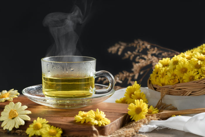 Steam rises from a glass cup of Chrysanthemum tea, surrounded by vibrant yellow Chrysanthemum flowers, evoking a sense of warmth and natural relaxation.