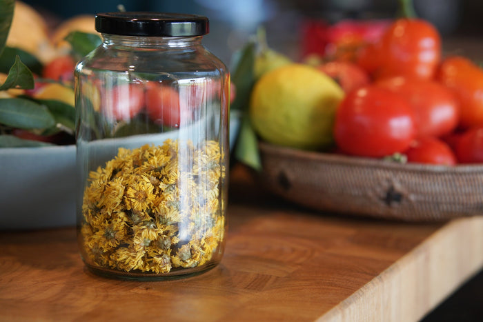 A glass jar filled with dried chrysanthemum flowers sits on a wooden kitchen counter. In the background, a woven basket holds fresh tomatoes and lemons, adding a vibrant contrast to the earthy tones of the dried flowers.