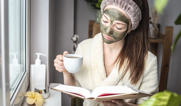 A woman wearing a green facial mask and a fluffy bathrobe, relaxing with a cup of tea and a book by the window, creating a self-care and skincare routine atmosphere.