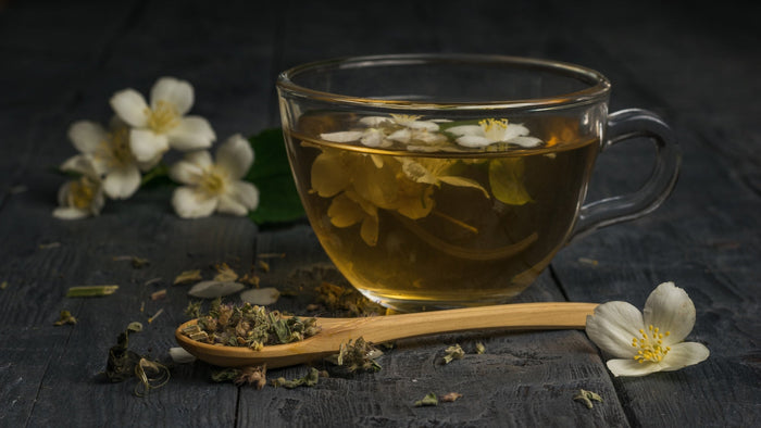 A glass cup of freshly brewed jasmine tea with floating jasmine flowers, placed on a dark wooden surface, accompanied by dried tea leaves on a wooden spoon and fresh jasmine blossoms in the background.