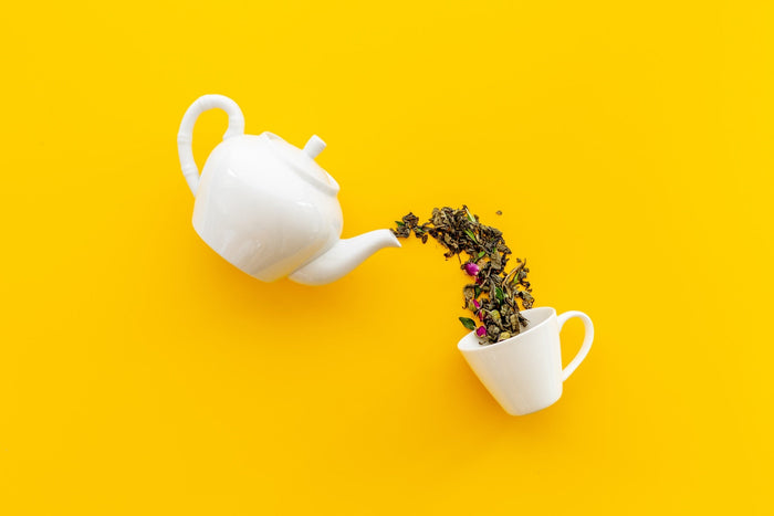 A white teapot appears to be pouring loose-leaf tea with rosebuds into a matching white teacup against a vibrant yellow background. 