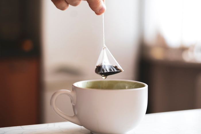 A close-up of a hand lifting a pyramid-shaped tea bag from a white ceramic cup, with a drop of tea falling, highlighting the brewing process of premium loose-leaf tea bags.