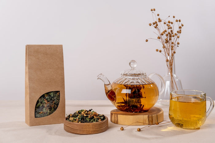 A glass teapot filled with golden brewed herbal tea on a wooden coaster, accompanied by a matching glass teacup, a kraft paper tea bag with a leaf-shaped window showing loose tea, and a wooden dish with dried tea leaves, set against a minimalist beige bac