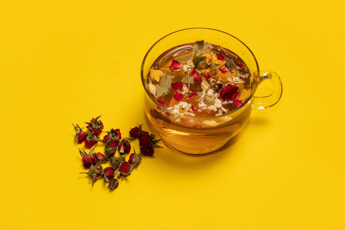A glass cup of herbal flower tea infused with dried rosebuds, chamomile, and other botanicals, placed on a vibrant yellow background. A small pile of dried rosebuds rests beside the cup, adding to the aesthetic appeal.