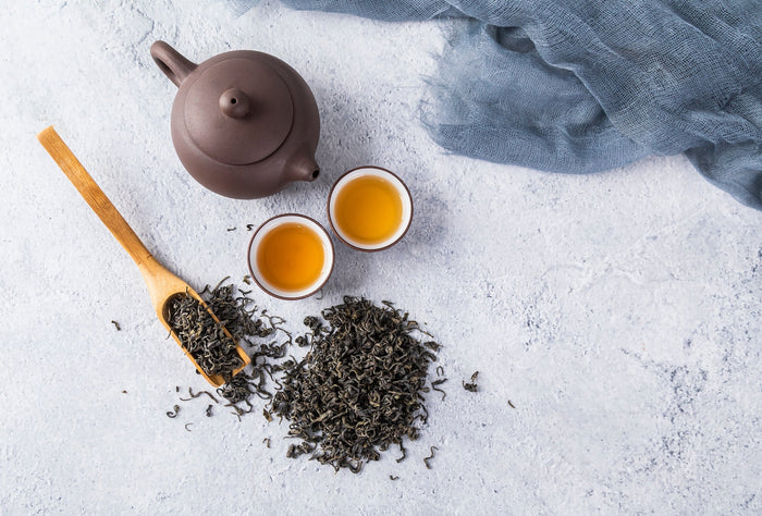 Flat lay of a teapot, two cups of golden tea, and loose tea leaves on a light textured background.
