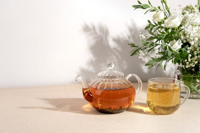 A glass teapot filled with amber-colored brewed tea alongside a matching glass teacup on a beige tablecloth, with a vase of white roses and greenery in the background, casting soft shadows on the wall.