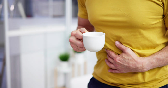 A man wearing a yellow shirt holding a white cup while clutching his stomach, possibly experiencing digestive discomfort.