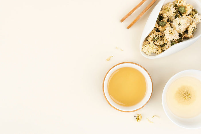 A minimalist top-down view of a freshly brewed cup of chrysanthemum tea with a golden hue, accompanied by a bowl of dried chinese chrysanthemum tea flowers and wooden chopsticks on a neutral background.