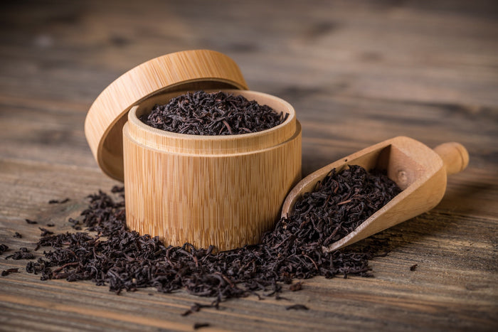 A wooden container filled with loose Lapsang Souchong black tea leaves, with a wooden scoop beside it, spilling tea leaves onto a rustic wooden surface. The deep, dark tea leaves contrast with the natural wood tones, emphasizing the rich, smoky character 