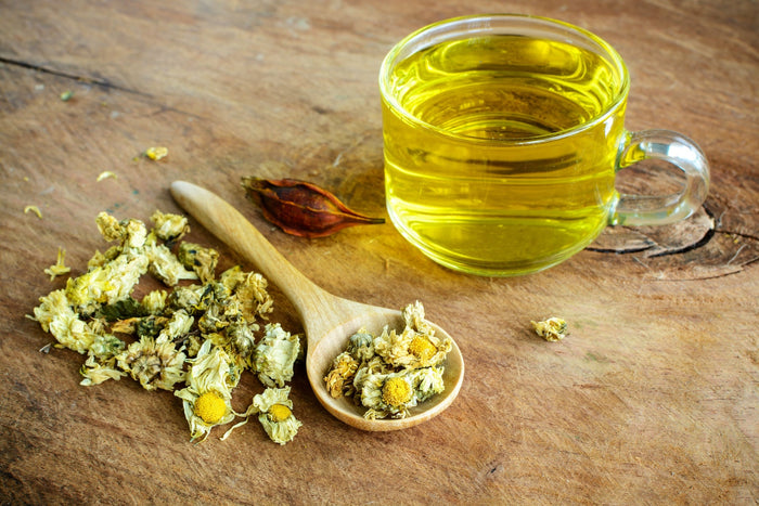 A glass cup filled with golden chrysanthemum tea sits on a rustic wooden surface, next to a wooden spoon holding dried chrysanthemum flowers.
