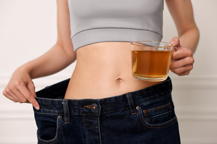 A fit woman wearing a gray sports top and loose-fitting jeans, holding a cup of tea in one hand while pulling the waistband of her jeans to show weight loss.