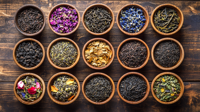 A top-down view of wooden bowls filled with a variety of organic loose leaf teas and herbal blends, arranged on a rustic wooden surface. 