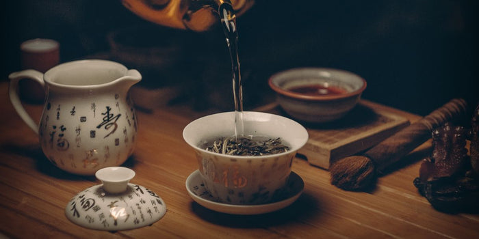 Traditional tea ceremony setup with hot water being poured into a bowl of loose tea leaves, surrounded by elegant Chinese teaware on a wooden table.