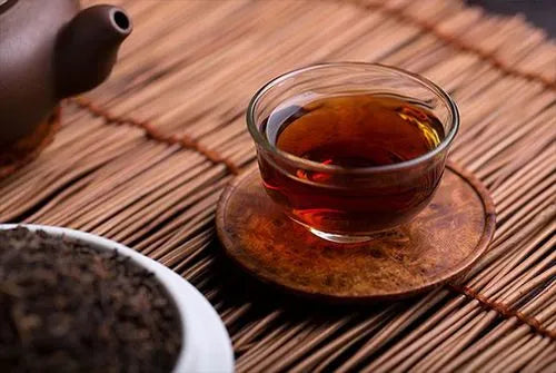 A glass cup filled with dark amber Da Hong Pao Oolong tea placed on a wooden coaster, with loose tea leaves in a white bowl and a teapot in the background, all set on a bamboo mat.